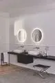Zwei runde Waschtische mit verchromten Armaturen von Ideal Standard an einer schwarzen Wandablage. Helle Wände und kugelrunde Spiegel mit indirekter Beleuchtung.